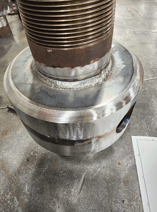 Close-up of heavy threaded shaft welded to a machined steel base in a fabrication shop.