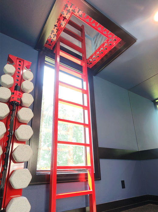 Red powder-coated steel ladder leading to a ceiling hatch framed with perforated cross-pattern panels in a gym.