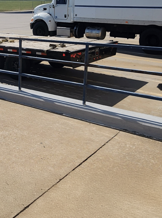 Steel safety railing installed along concrete loading area beside a flatbed trailer and box truck.