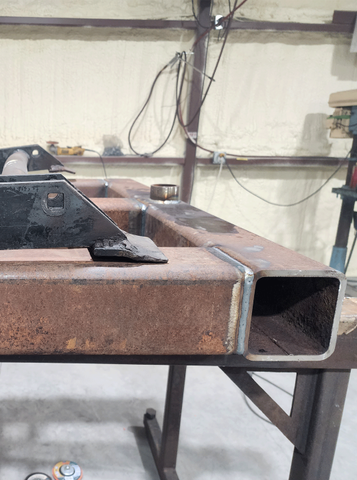 End view of rectangular steel tubing with welded seams and bracket on a fabrication table.