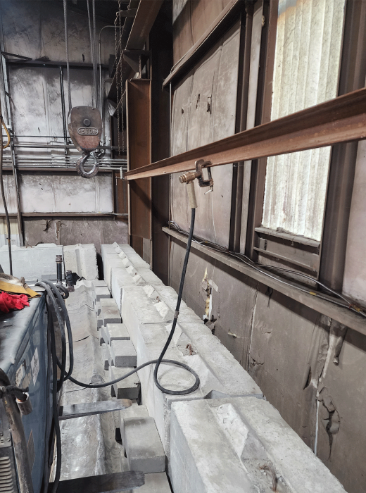 Industrial shop interior with stacked concrete blocks, overhead hoist, and welding leads.