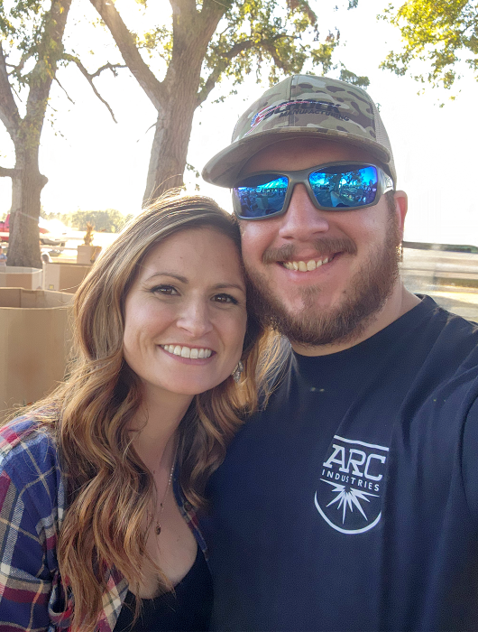 Smiling couple standing outdoors in front of trees, with the man wearing an ARC Industries shirt and cap.