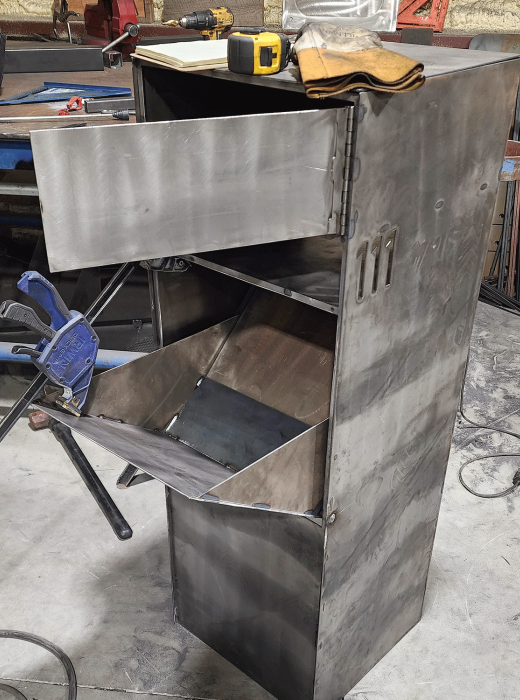 Custom fabricated steel bin cabinet with hinged chute doors open in a metal shop.